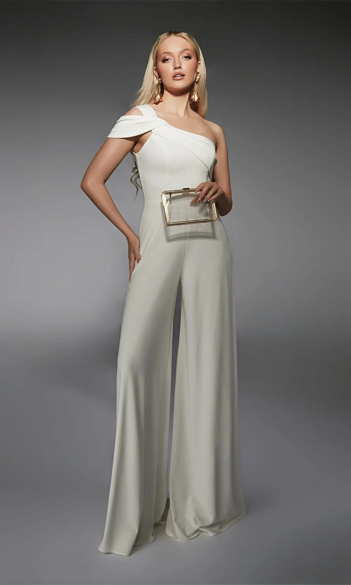 A person stands in an ivory Long Formal Jumpsuit: Alyce 70066 with wide-leg pants, holding a transparent box clutch against a gray background.